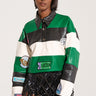 A person stands against a plain background, wearing the BIGGEST FAN LONG SLEEVE RUGBY TOP, which features green, white, and black striped color block design with embroidered letterman patches and a black collar. They also have on a black geometric skirt made of diamond-shaped pieces. Their hair is straight and shoulder-length.