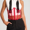 A woman stands with one hand in her pocket, wearing a unique outfit featuring a sleeveless white shirt with black panels and the MESH BELT - a red mesh corset with a back lace-up panel overlaying it. She pairs it with black pinstripe pants.