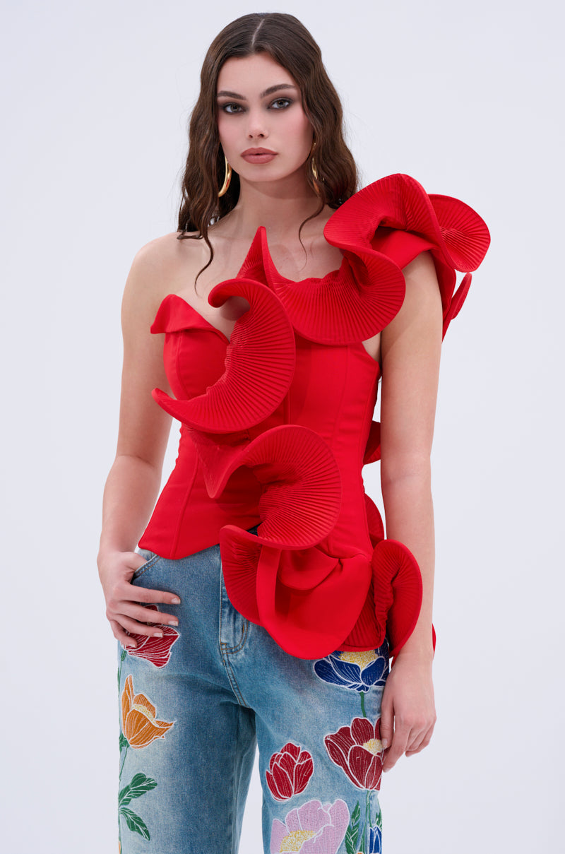 A woman with wavy brown hair and gold hoop earrings wears the ABSTRACT RUFFLE OFF THE SHOULDER BLOUSE in bold red, paired with blue jeans featuring vibrant floral embroidery, set against a plain light background.