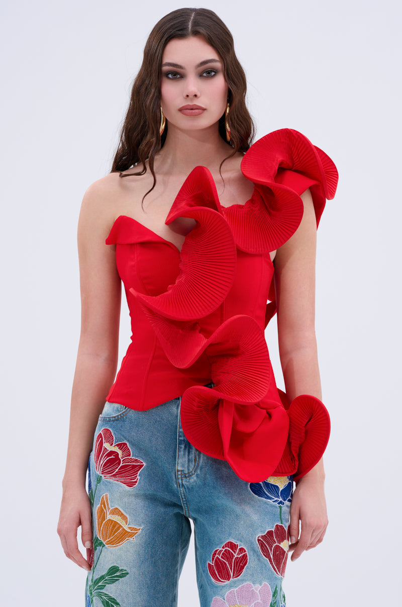 A woman with long brown hair poses against a plain light background wearing the ABSTRACT RUFFLE OFF THE SHOULDER BLOUSE in bright red with sculptural ruffles and blue jeans embellished with colorful embroidered flowers.