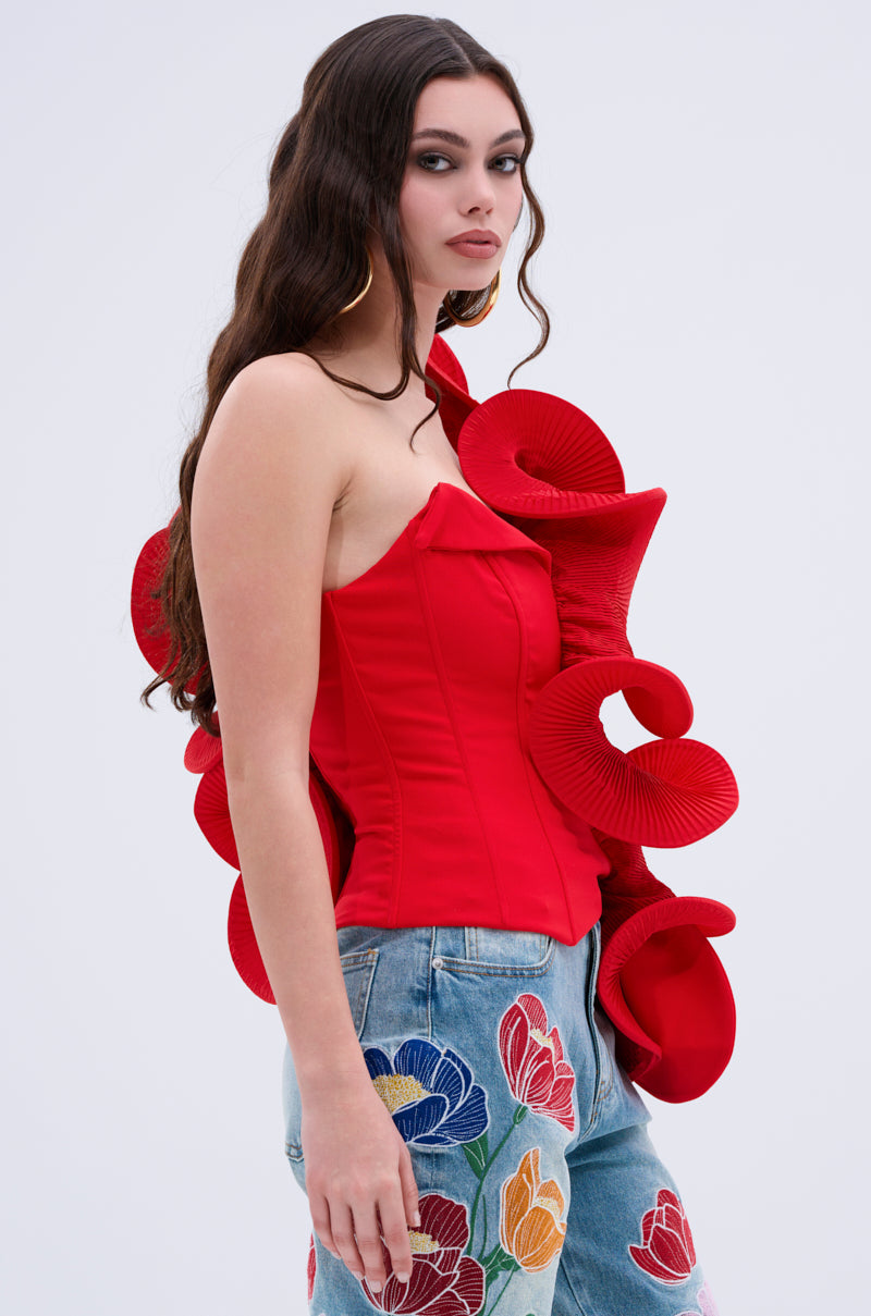 A woman with long wavy brown hair models the ABSTRACT RUFFLE OFF THE SHOULDER BLOUSE in bold red, featuring dramatic sculpted ruffles. She pairs it with light blue jeans with colorful floral embroidery, set against a plain white background.
