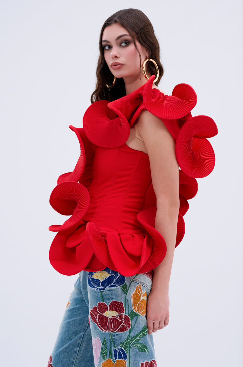 A woman with long, wavy brown hair models the ABSTRACT RUFFLE OFF THE SHOULDER BLOUSE—an eye-catching red top with bold ruffled details—paired with blue jeans featuring colorful embroidered florals against a plain light background.