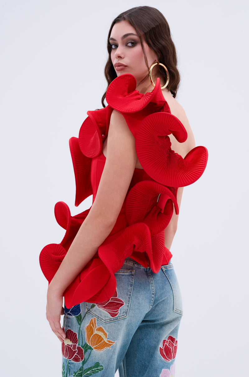 A woman with long brown hair wears the ABSTRACT RUFFLE OFF THE SHOULDER BLOUSE in red and large gold hoop earrings, paired with light blue floral-embroidered jeans, set against a plain light gray background.