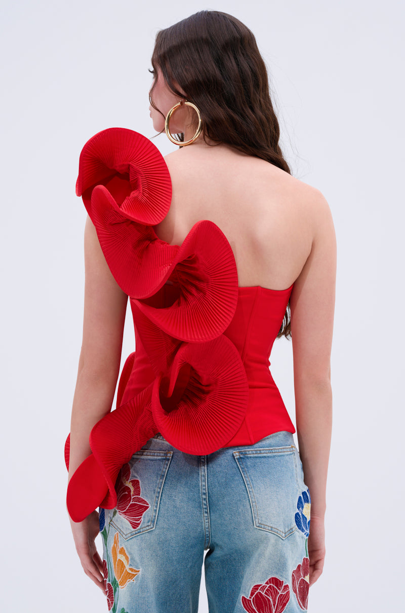 A woman with long, wavy brown hair wears the ABSTRACT RUFFLE OFF THE SHOULDER BLOUSE in red, styled with blue jeans featuring colorful floral pocket embroidery. She faces away from the camera against a plain background.