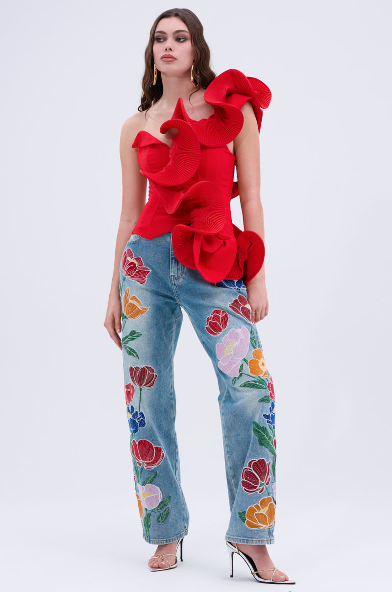 A woman with long wavy hair wears the ABSTRACT RUFFLE OFF THE SHOULDER BLOUSE in bright red, paired with blue jeans featuring colorful floral embroidery and clear high-heeled shoes, standing against a plain light background.