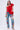 A woman with long wavy hair wears the ABSTRACT RUFFLE OFF THE SHOULDER BLOUSE in bright red, paired with blue jeans featuring colorful floral embroidery and clear high-heeled shoes, standing against a plain light background.