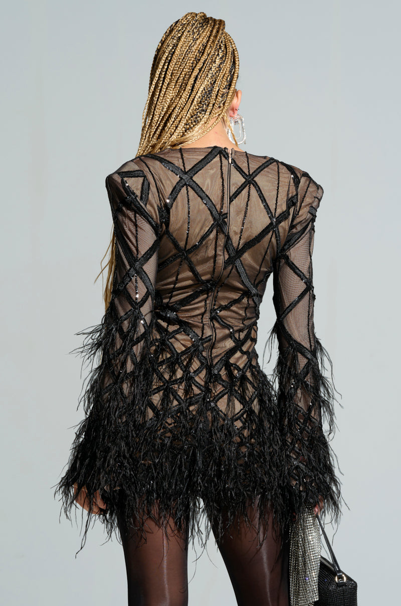 A person with long braided hair is wearing the SHOW ME A GOOD TIME MESH MINI DRESS, adorned with geometric patterns and feather detailing. They are facing away, holding a handbag, and large hoop earrings are visible against a neutral gray background.