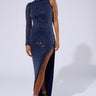 A woman stands posing in the Ulyssa Rhinestone Mesh Maxi Dress in navy blue, showcasing its stretchy mesh fabrication. The dress features an asymmetrical single-sleeve silhouette adorned with small, sparkling rhinestones and a high slit on one side that reveals her leg. She completes the look with strappy high heels and has her hair pulled back tightly. The background is plain and light-colored.