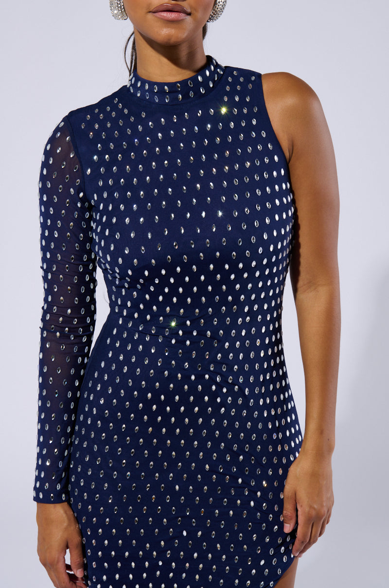 A person wearing the Ulyssa Rhinestone Mesh Maxi Dress, characterized by its asymmetrical single-sleeve silhouette with one long sleeve and one sleeveless side. The dress is navy blue and fitted, adorned with numerous small silver studs. Made from stretchy mesh fabrication, the background is plain and light-colored, their face partially visible.