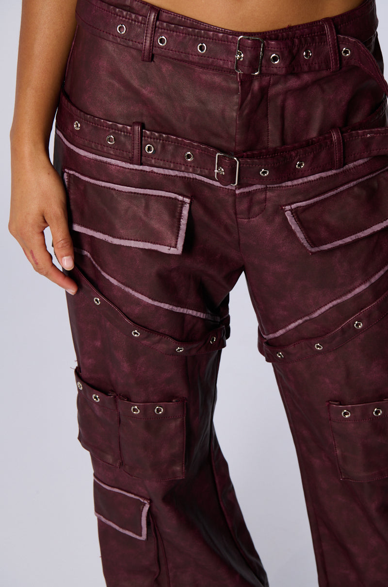 A person wearing the stylish DESERT VIBE CARGO PANT IN BURGUNDY, featuring a high-rise fit with multiple pockets, belts, and metal eyelet details. The pants have a worn, textured appearance that adds an edgy look. The person stands with their left hand resting casually by their side.