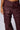 A person wearing the stylish DESERT VIBE CARGO PANT IN BURGUNDY, featuring a high-rise fit with multiple pockets, belts, and metal eyelet details. The pants have a worn, textured appearance that adds an edgy look. The person stands with their left hand resting casually by their side.