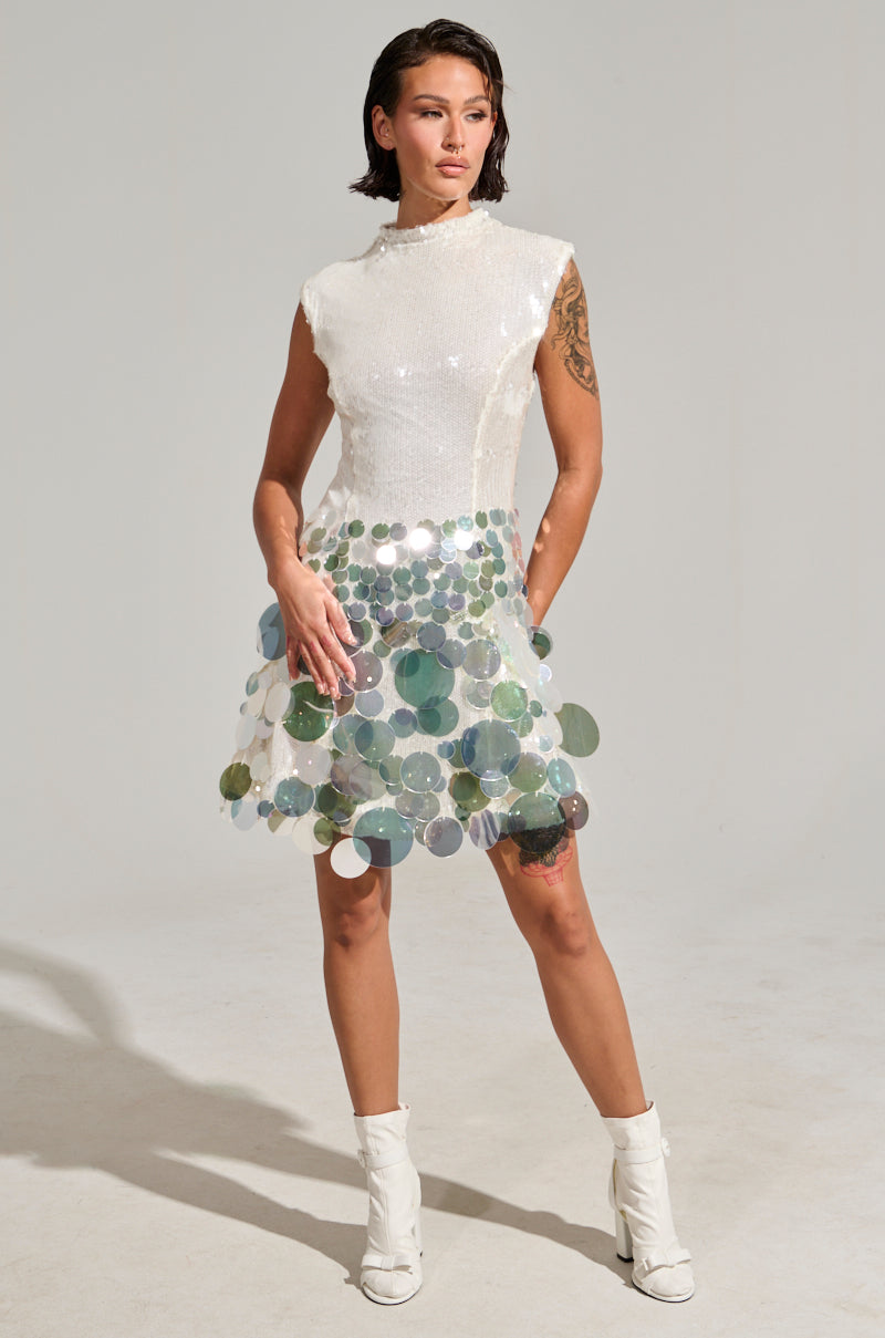 Dressed in the "All the Right Ways" sequin mini dress, a person exudes glam with its shimmering bodycon silhouette. They pair it with white ankle boots and reveal a tattoo on their right arm against a plain light gray background.