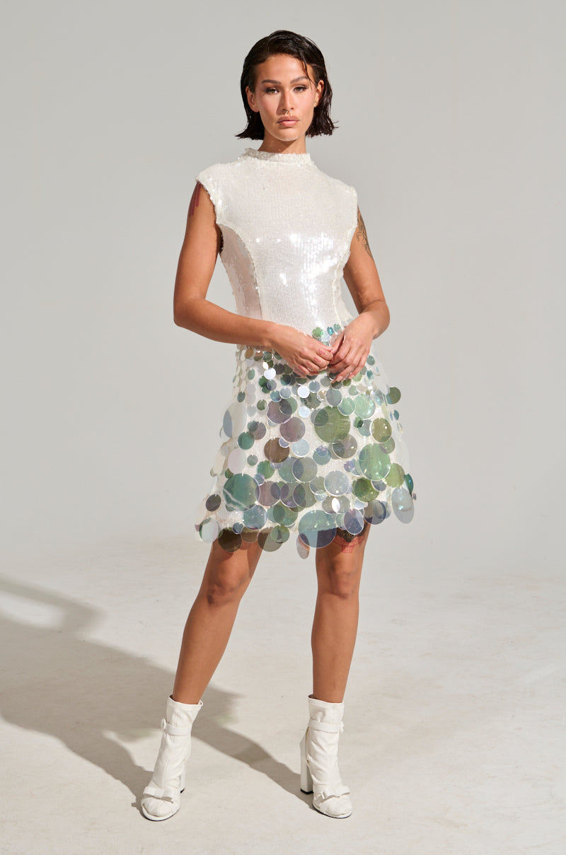 A person stands confidently against a plain background, wearing the ALL THE RIGHT WAYS SEQUIN MINI DRESS, which features a fitted white top paired with a skirt embellished with large translucent blue and green sequins. Their outfit is completed with stylish white ankle boots.