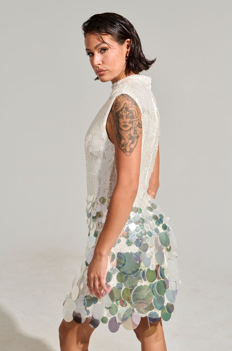 A woman with wet hair poses confidently in the ALL THE RIGHT WAYS SEQUIN MINI DRESS, which is sleeveless and semi-transparent, perfectly accentuating her bodycon silhouette. Her arm showcases a prominent tattoo against the plain, light gray background, exuding an effortlessly chic party vibe.