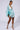 A woman stands posing in the LIFE'S A BREEZE MESH MINI DRESS WITH RHINESTONES, a light blue shimmering draped dress featuring a deep plunging neckline and batwing sleeves. She is wearing high-heeled, strappy sandals and large hoop earrings against a plain white background.