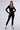 A person stands confidently against a plain background, wearing the HOT GIRL WALK LONG SLEEVE CATSUIT IN BLACK paired with black and white sneakers. They accessorize with large hoop earrings, a necklace, and have a small tattoo visible on their chest.