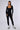 A person stands confidently against a plain background wearing the HOT GIRL WALK LONG SLEEVE CATSUIT IN BLACK, a sleek and fitted ribbed knit piece featuring long sleeves and a square neckline. They accessorize with hoop earrings, a chain necklace, and white sneakers with black accents. One hand is tucked into a pocket while the other rests on their thigh.