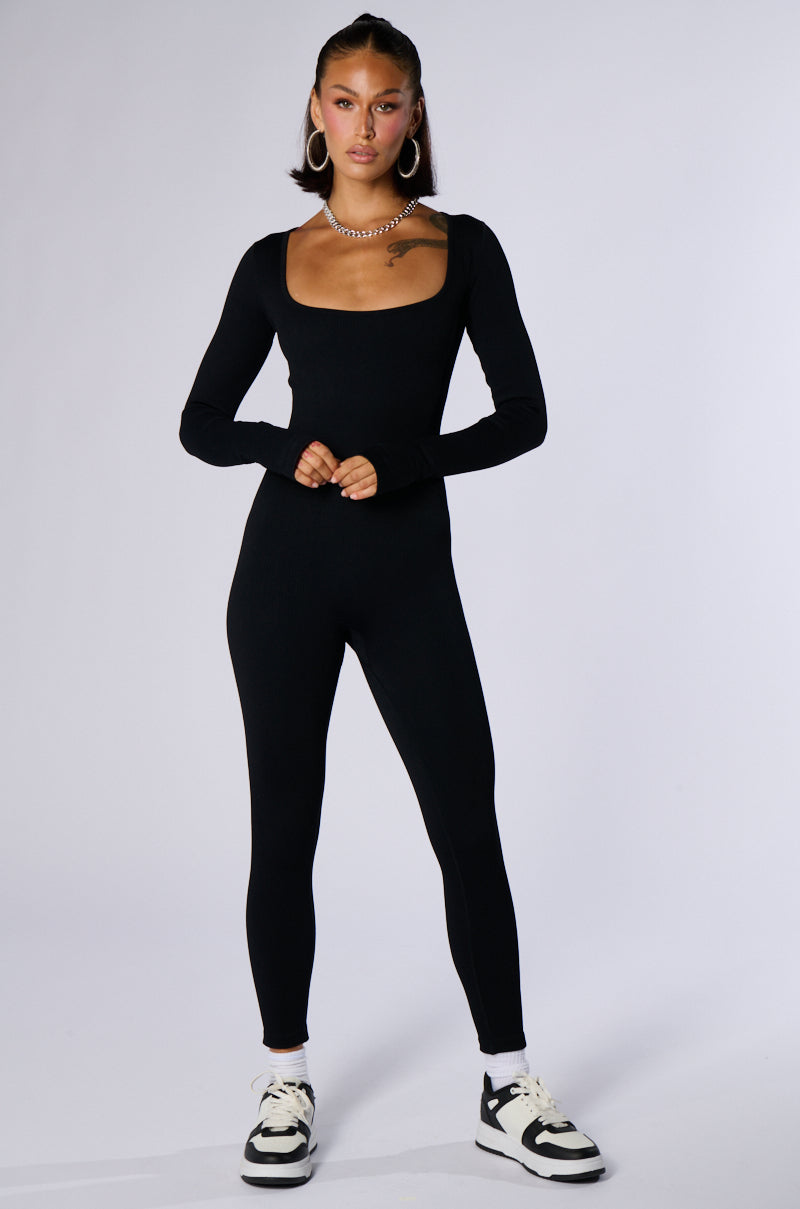 A person with long hair stands confidently against a plain background, wearing the HOT GIRL WALK LONG SLEEVE CATSUIT IN BLACK, white and black sneakers, hoop earrings, and a necklace. Their hands are held together at their waist.