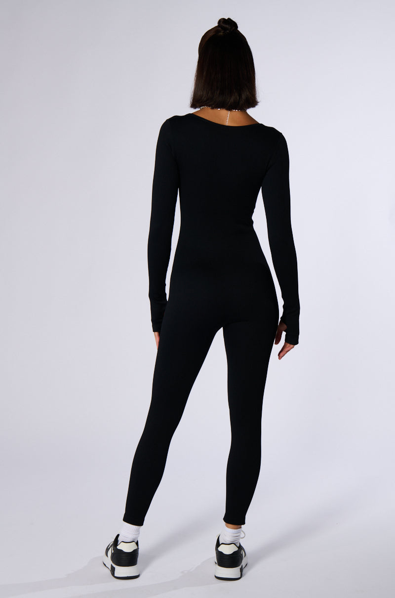A person wearing the HOT GIRL WALK LONG SLEEVE CATSUIT IN BLACK and black-and-white athletic shoes stands facing away, hands relaxed by their sides against a plain white background.