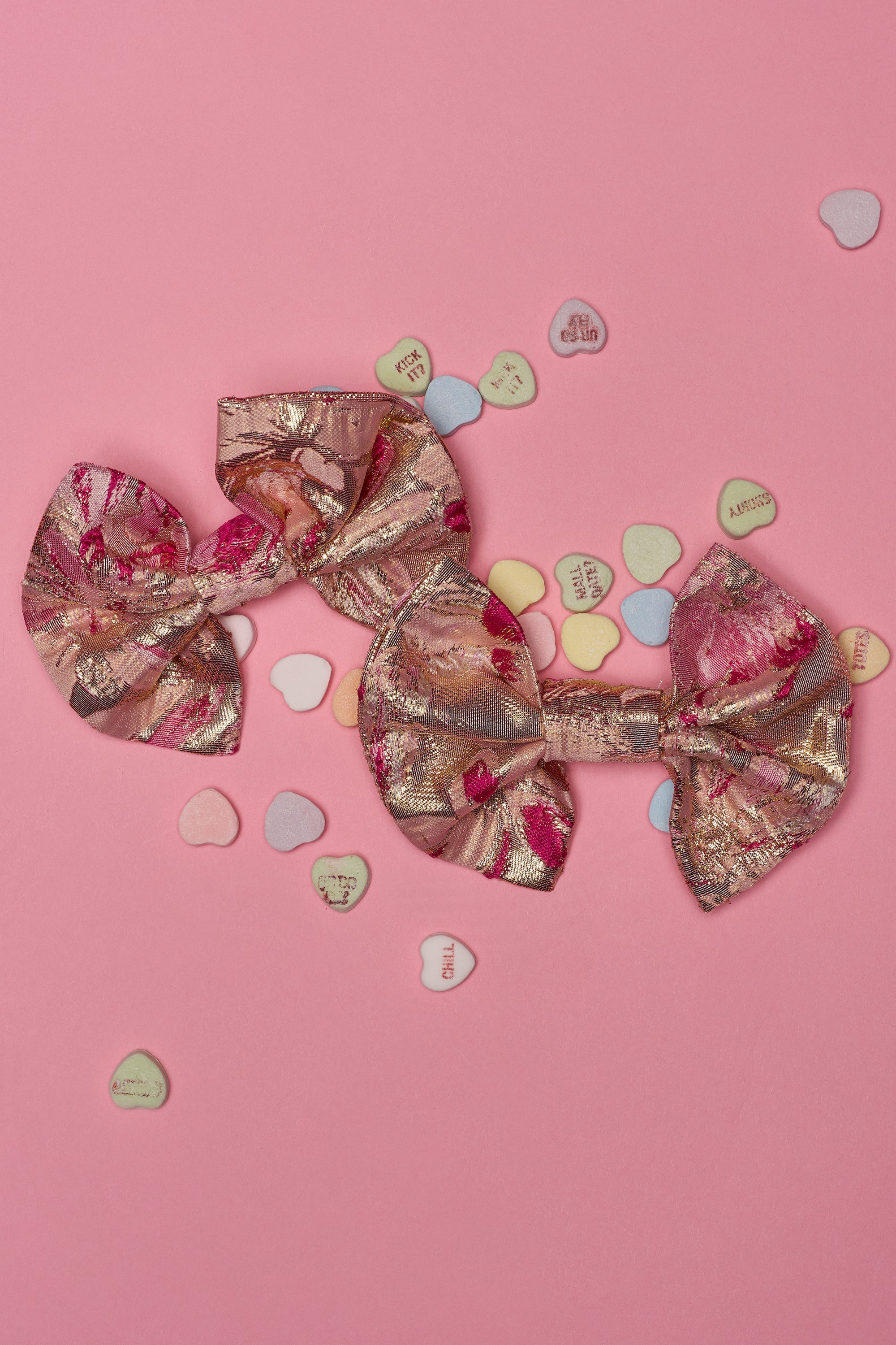 Two rose gold floral hair bows rest on a pink surface with pastel candy hearts, perfectly paired with the REALLY ME BOW EARRINGS for a charming, romantic look. 0