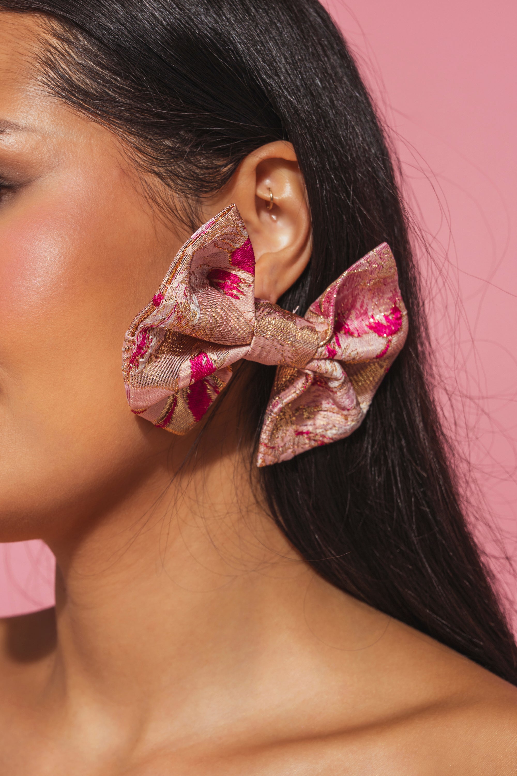 A close-up of a woman with straight dark hair wearing REALLY ME BOW EARRINGS—large, shiny pink and gold brocade bows positioned above her ear—set against a pink background. 1