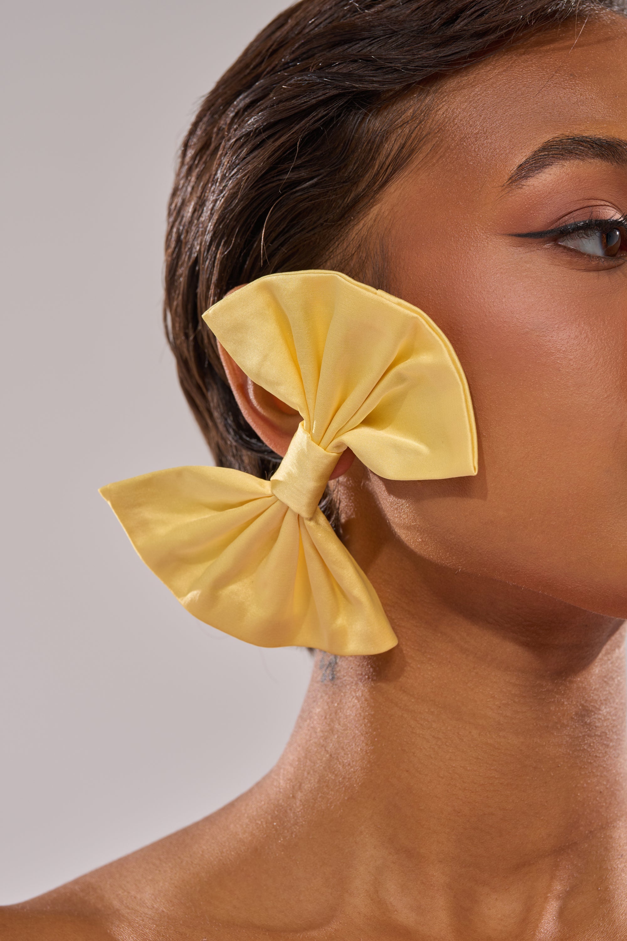Close-up of a person with short brown hair wearing SPRING SZN BOW EARRINGS—oversized, yellow statement pieces that cover most of the ear, set against a plain, light background. 2