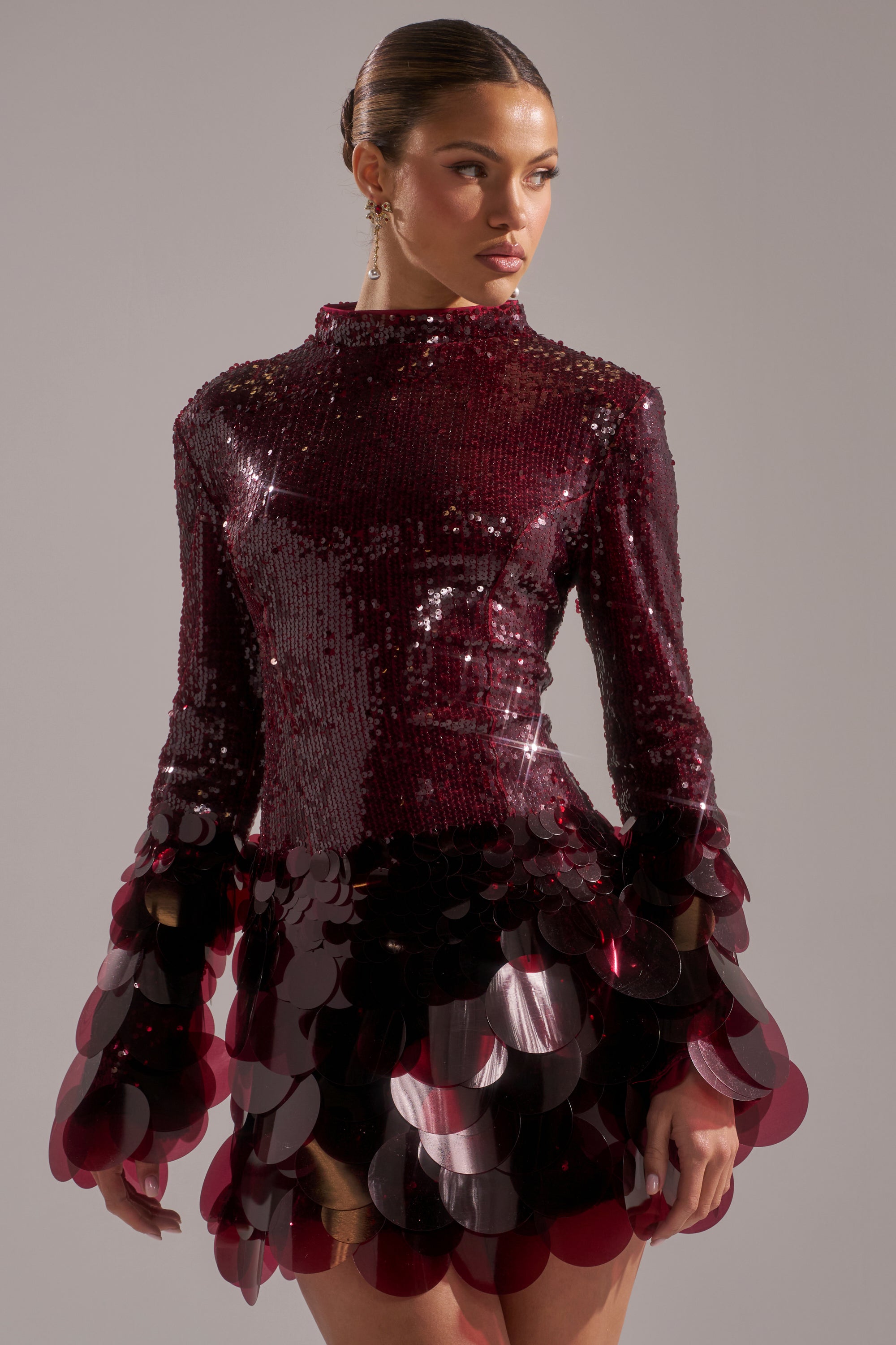 A woman models THE MOMENT SEQUIN MINI DRESS IN BURGUNDY, featuring large circular sequin details on the sleeves and skirt, standing against a plain light background and gazing slightly to the side.