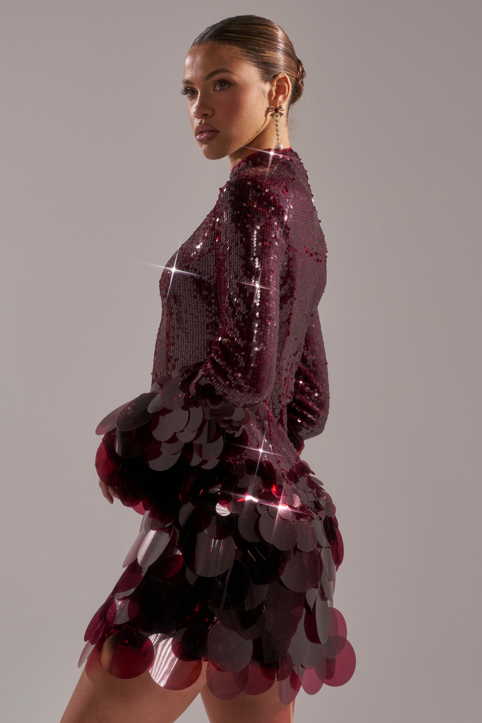 A woman stands in profile against a plain gray background, wearing THE MOMENT SEQUIN MINI DRESS IN BURGUNDY, adorned with sparkling sequins and large shiny circular embellishments on the skirt and sleeves.