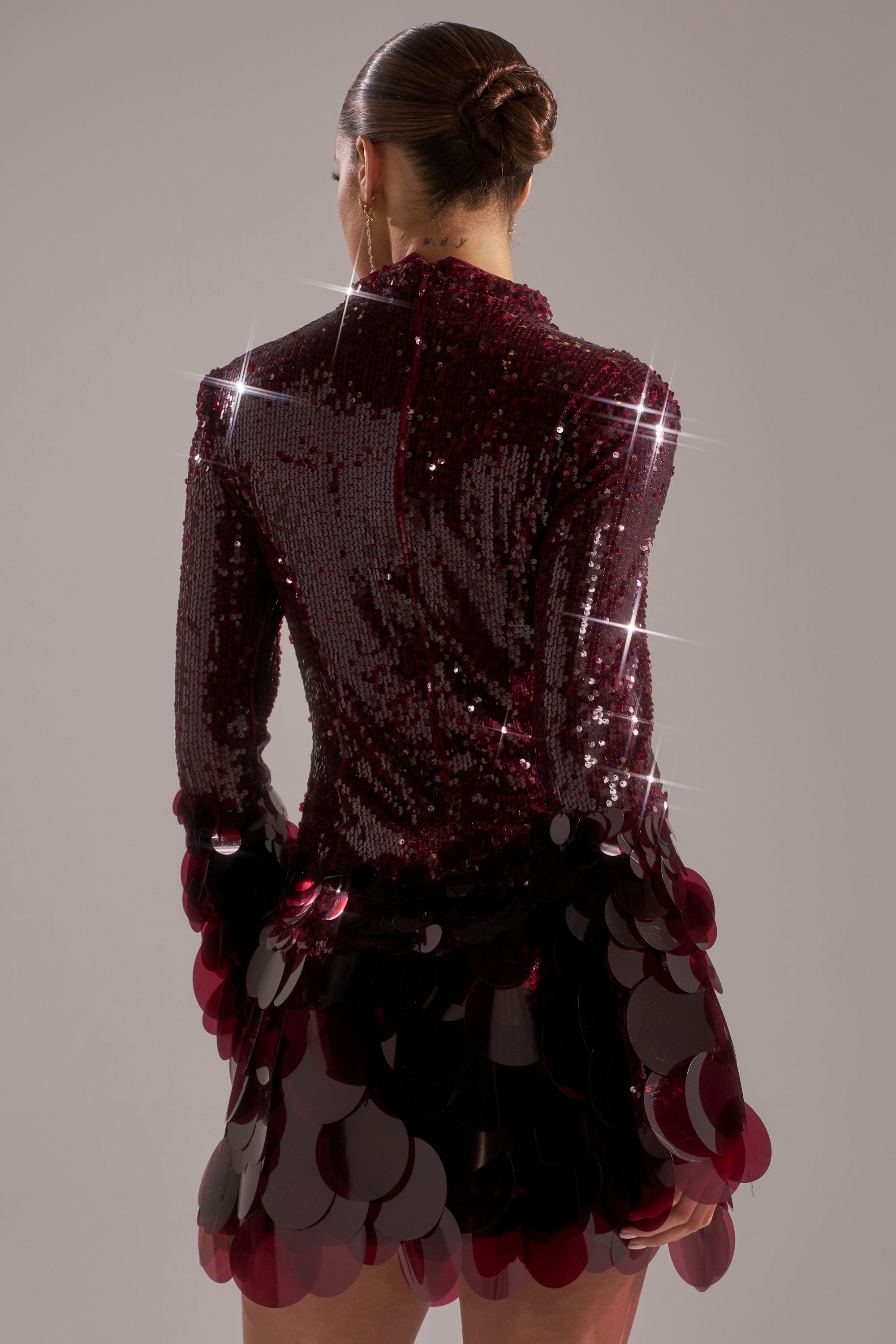A woman in a sleek bun wears THE MOMENT SEQUIN MINI DRESS IN BURGUNDY, featuring sparkling circular sequin embellishments on the sleeves and hem, as she stands with her back to the camera against a plain background.