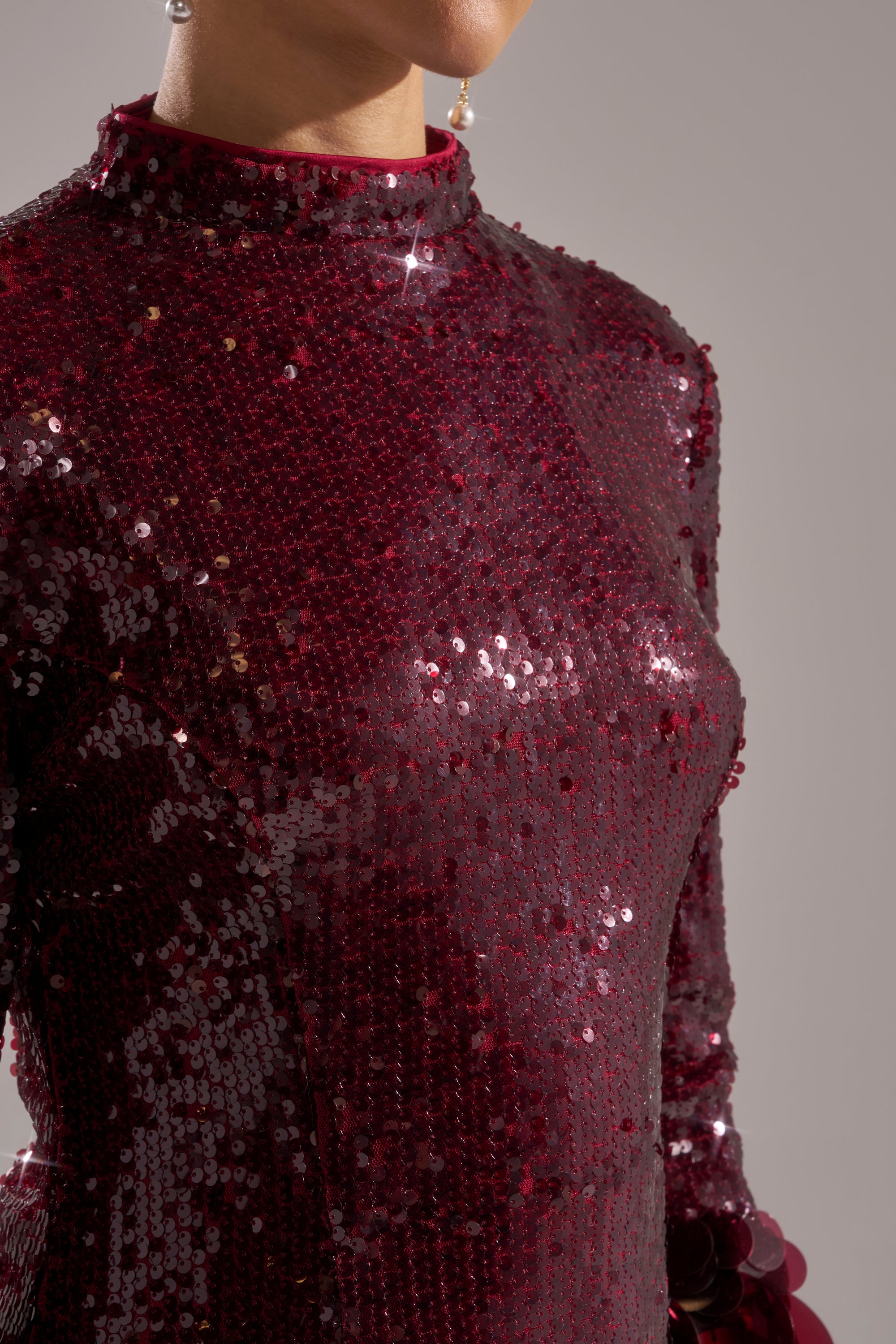 A person showcases THE MOMENT SEQUIN MINI DRESS IN BURGUNDY, a high-neck, long-sleeve mini dress with sparkling sequins. Only their lower face, neck, shoulder, and upper body are visible against a plain neutral background.