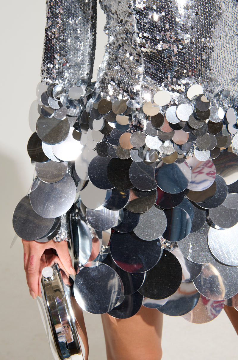 Wearing THE MOMENT SEQUIN MINI DRESS, adorned with large reflective discs, a person holds a silver clutch. The close-up showcases the shimmering textures and intricate sequin details of the bodycon style against a plain background.