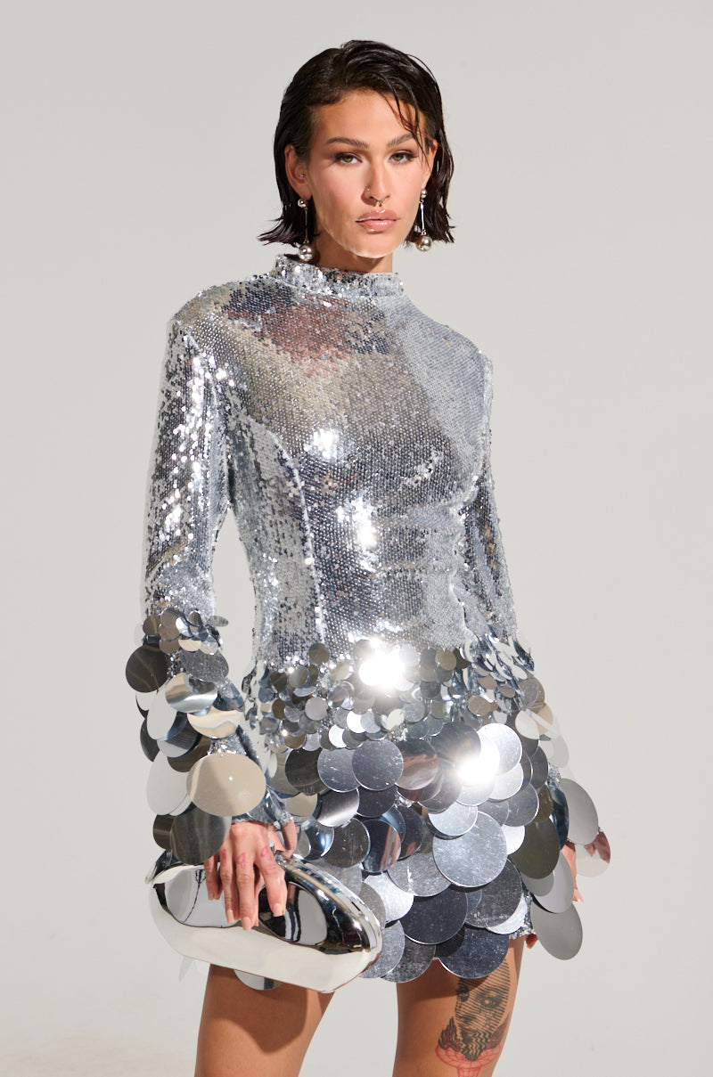 A person wearing THE MOMENT SEQUIN MINI DRESS, adorned with reflective sequin detailing, poses confidently. They hold a metallic clutch and have short, slicked-back hair, standing against a plain background.