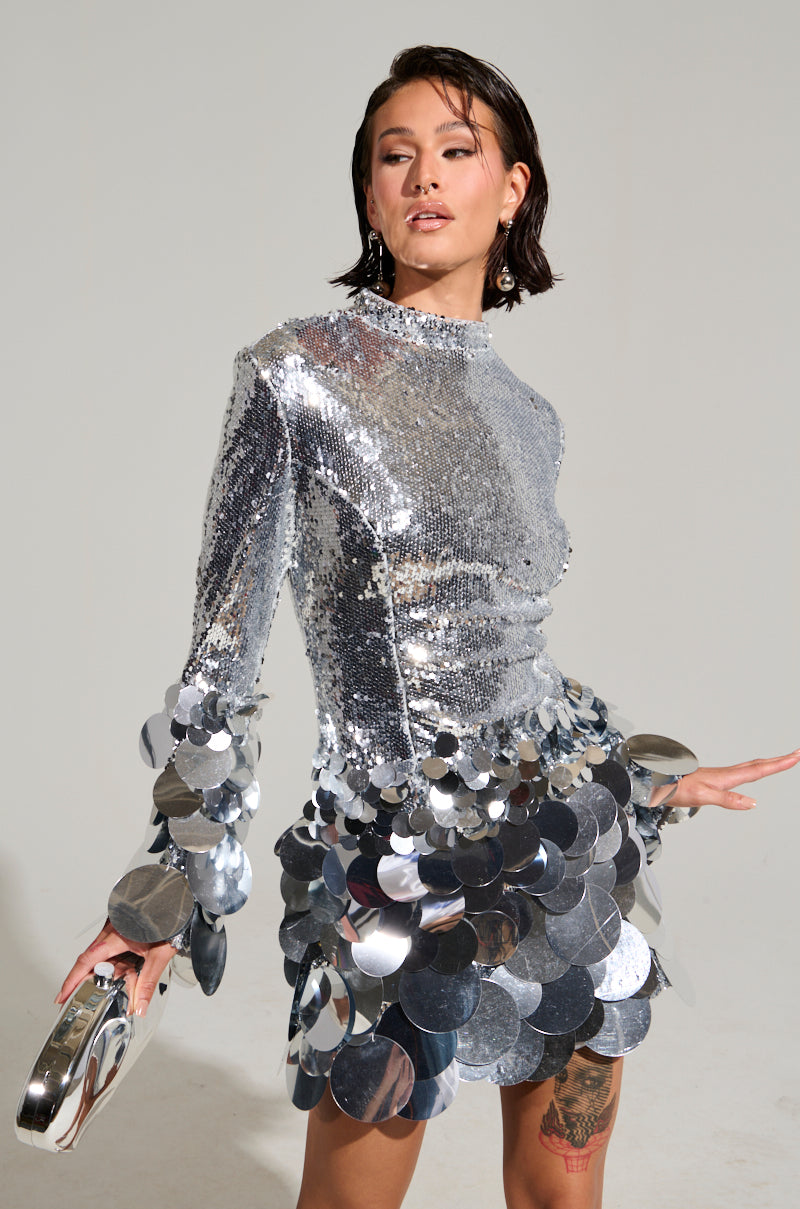 Wearing "THE MOMENT SEQUIN MINI DRESS," a person poses with a small silver handbag. The dress, a stunning bodycon style, features large circular sequin embellishments along with long sleeves and a high neckline, highlighted by reflective sequin detailing. They stand against a plain background.