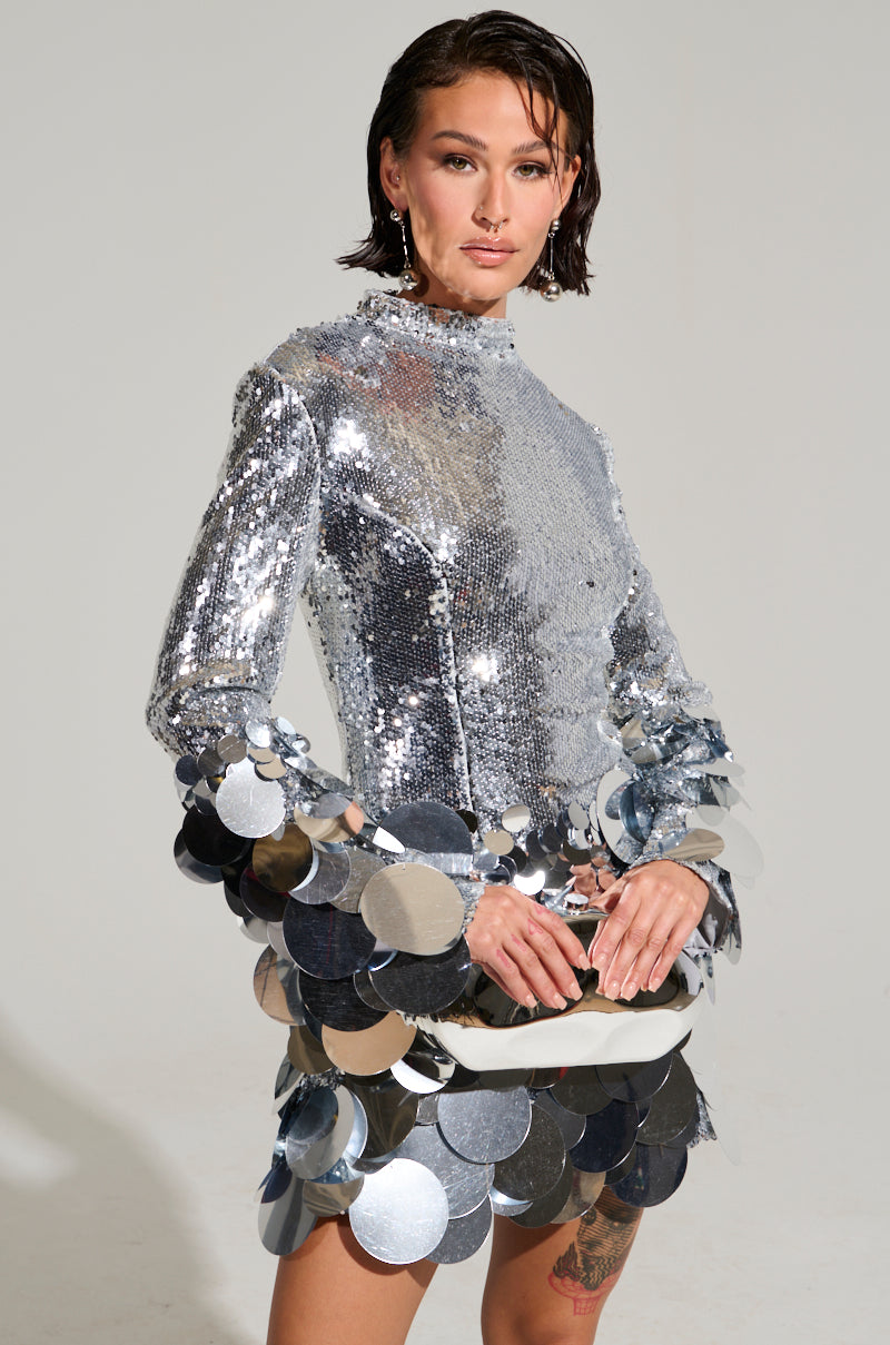 A person wearing "THE MOMENT SEQUIN MINI DRESS," which features a reflective silver finish, large circular embellishments, and a bodycon fit, stands against a plain background. The dress has long sleeves and a high neckline. They have short, dark hair and are posing confidently with hands clasped.