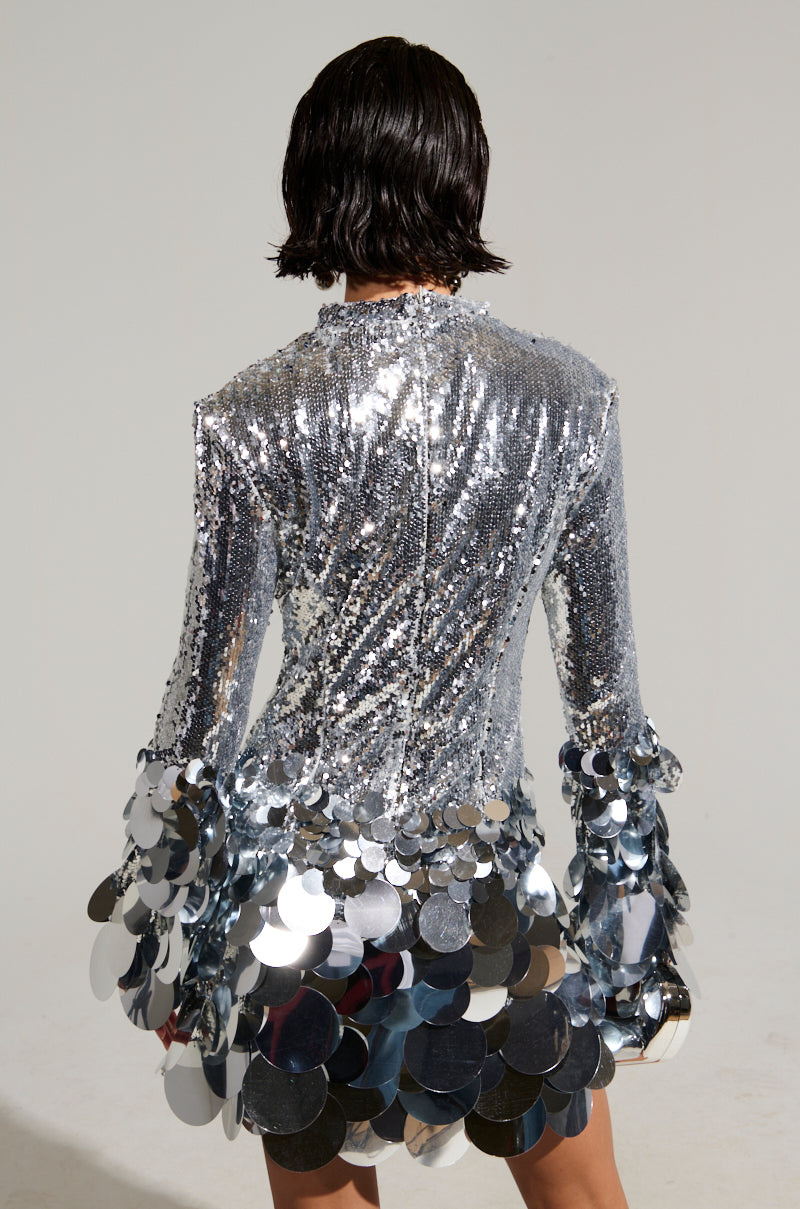 A person stands with their back to the camera, wearing THE MOMENT SEQUIN MINI DRESS, a shiny silver piece adorned with large circular embellishments at the hem and sleeves. The reflective sequin detailing catches the light brilliantly, creating a sparkling effect. The person has short, dark hair.