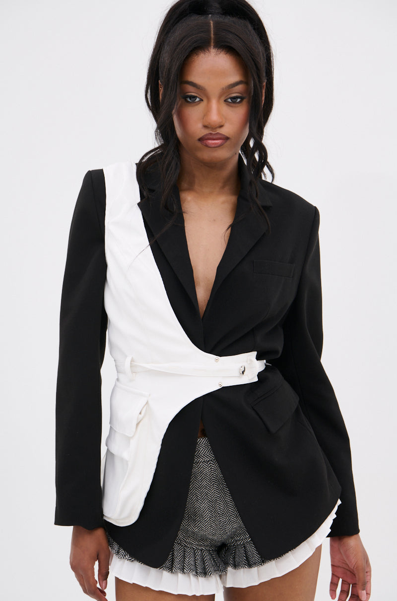 A woman with long dark hair wears a black blazer with an asymmetrical white overlay, textured shorts, pleated white trim, and the HERE FOR THE PLOT BELT. She stands against a plain light background, looking slightly downward.