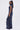 A woman with long straight hair in a high ponytail wears the DONT MESS WITH ME COLLARED DENIM MAXI DRESS, a sleeveless navy blue piece with utility pockets, standing sideways and looking at the camera.