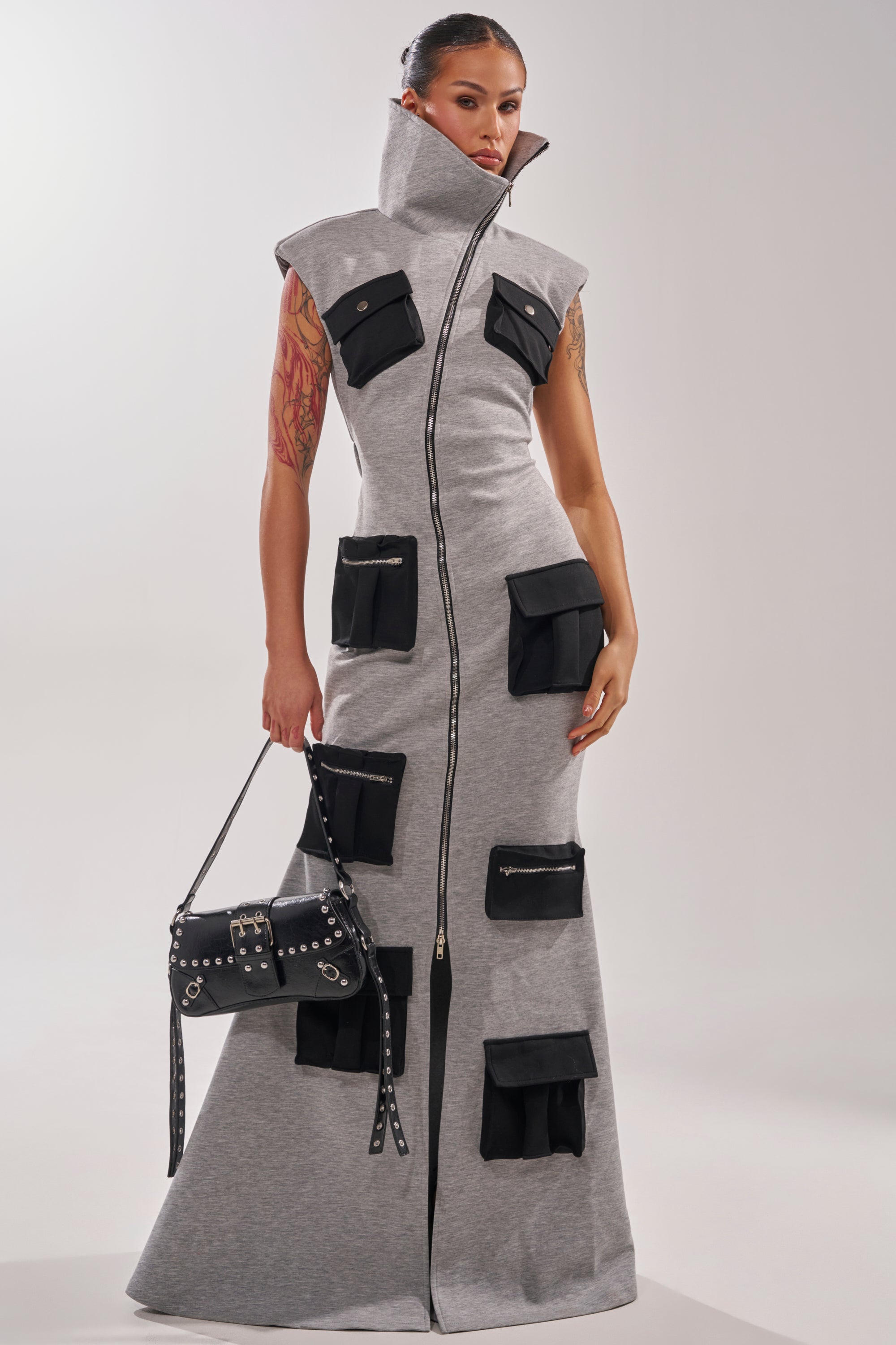A woman stands confidently, showing arm tattoos, in the DON'T MESS WITH ME COLLARED MAXI DRESS IN GREY SCUBA—a cargo-inspired, sleeveless maxi dress with a high collar and black pockets—holding a black studded handbag.