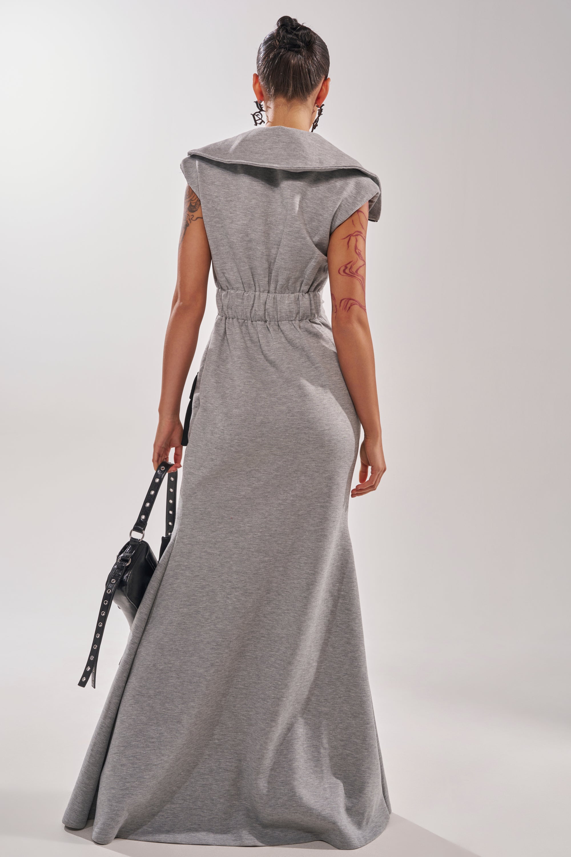 A woman with short hair faces away, wearing the DON'T MESS WITH ME COLLARED MAXI DRESS IN GREY SCUBA with a cinched waist and wide collar. She carries a black bag and displays tattoos on her right arm against a plain light background.