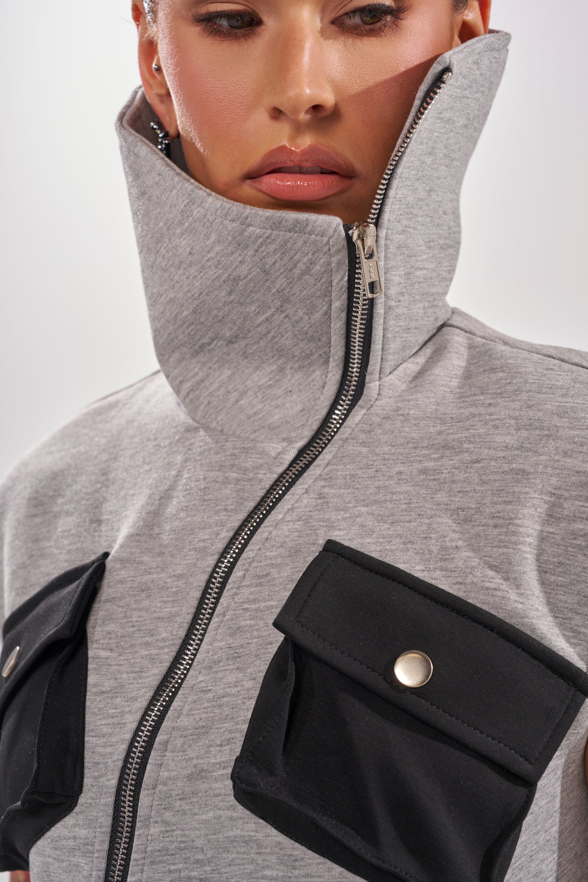 A woman wears the DON'T MESS WITH ME COLLARED MAXI DRESS IN GREY SCUBA, featuring a high collar, zip front, and large black pockets with silver buttons. Only the lower part of her face and upper torso are shown.