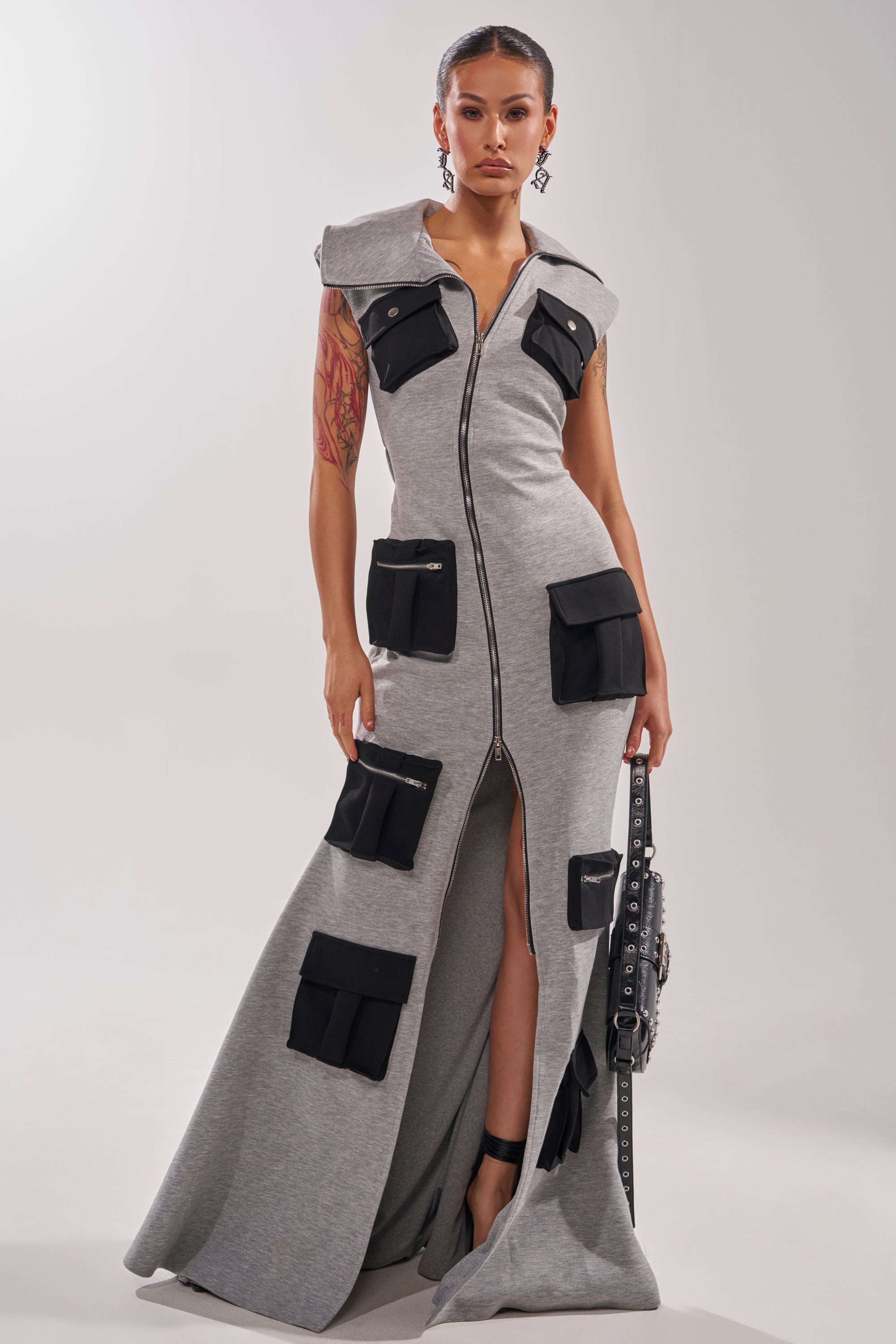 A woman with tattoos on one arm models the DON'T MESS WITH ME Collared Maxi Dress in grey scuba, featuring a zip front and multiple black pockets, while holding a black studded bag against a light background.