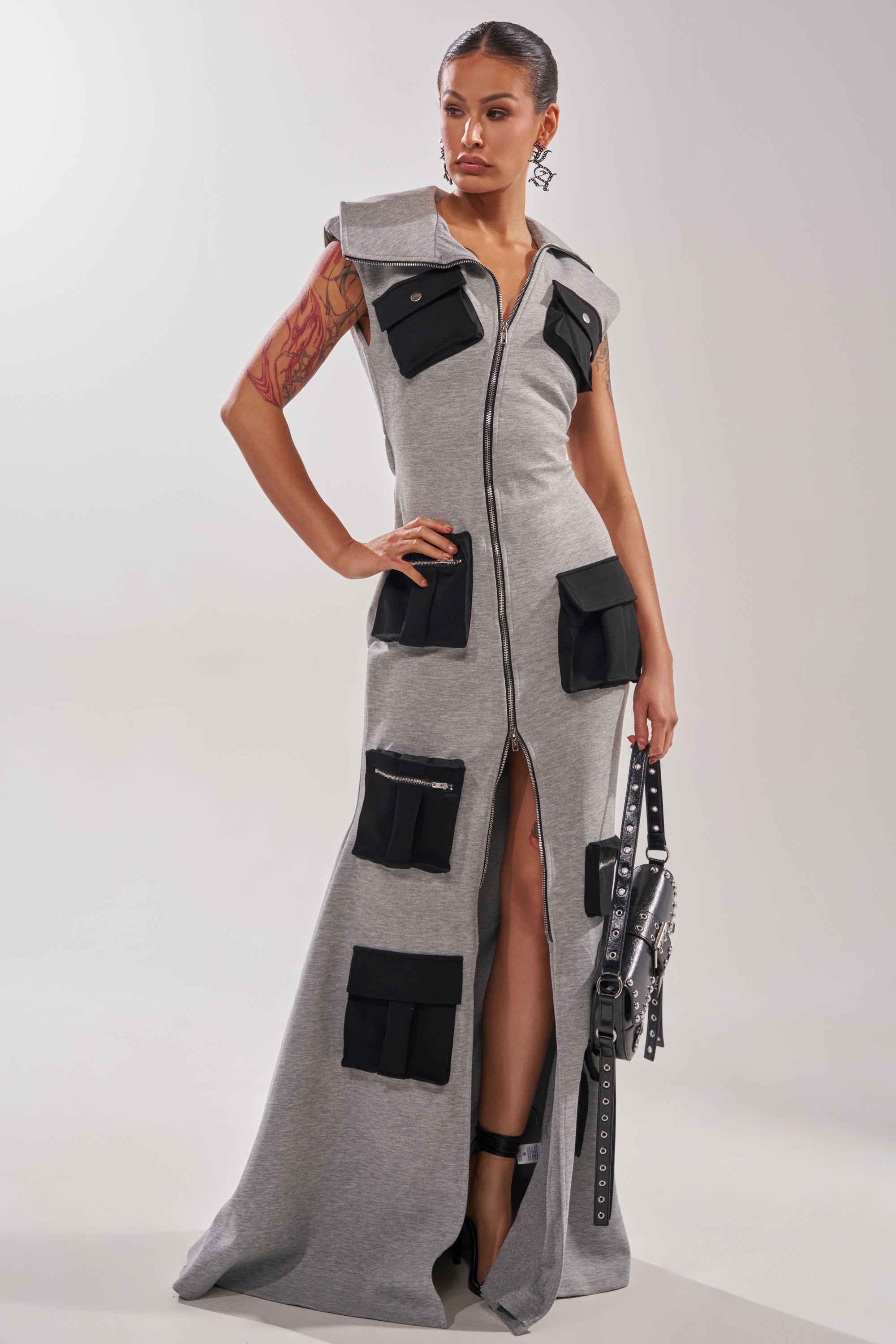 A woman stands confidently in the DON'T MESS WITH ME Collared Maxi Dress in Grey Scuba, a sleeveless utility style with a front zipper and black pockets. She carries a studded black handbag and has a tattoo on her upper right arm.