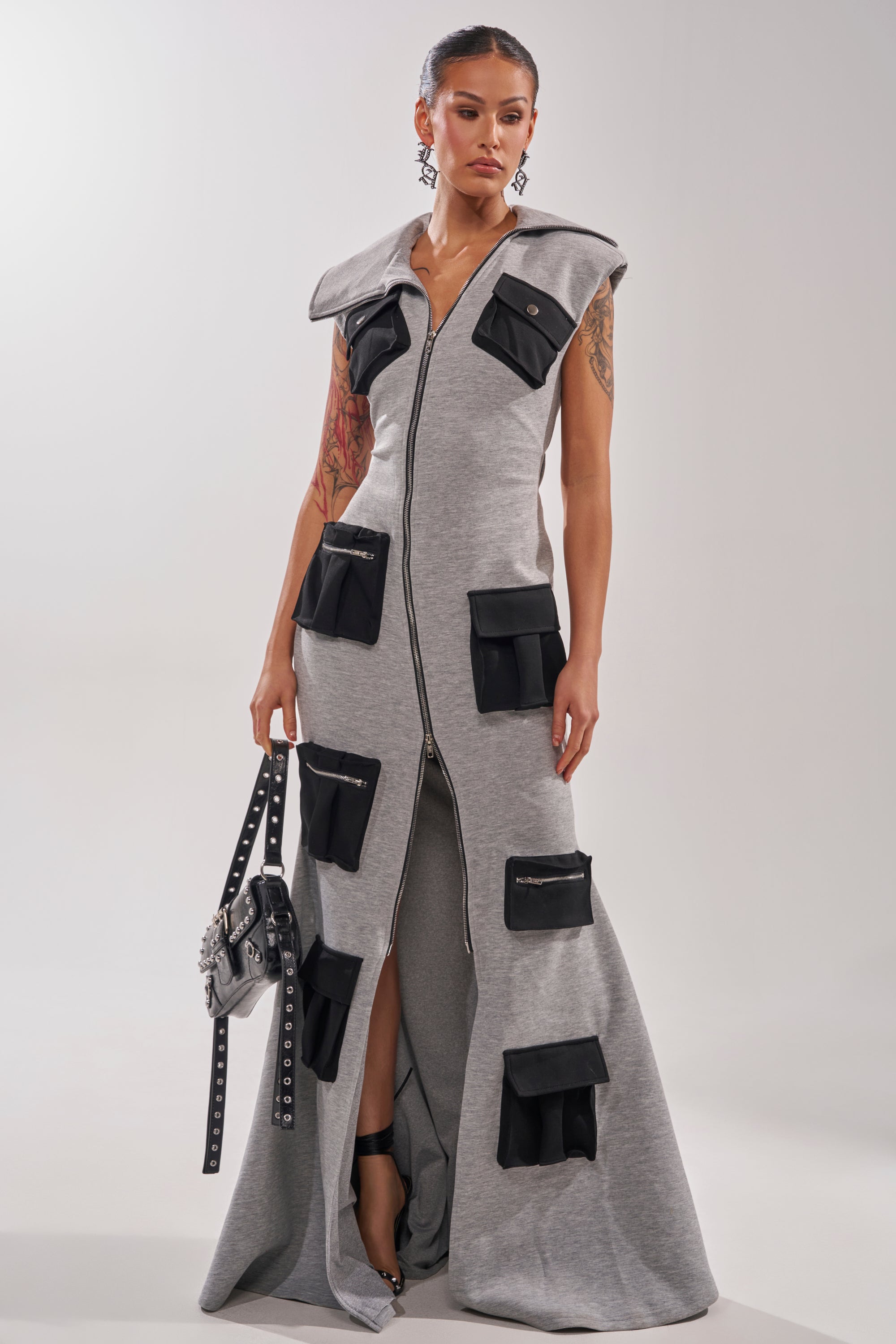 A woman in the DON'T MESS WITH ME Collared Maxi Dress in grey scuba—with black cargo pockets and a high slit—holds a studded handbag, standing against a plain background that highlights her earrings and tattoos.