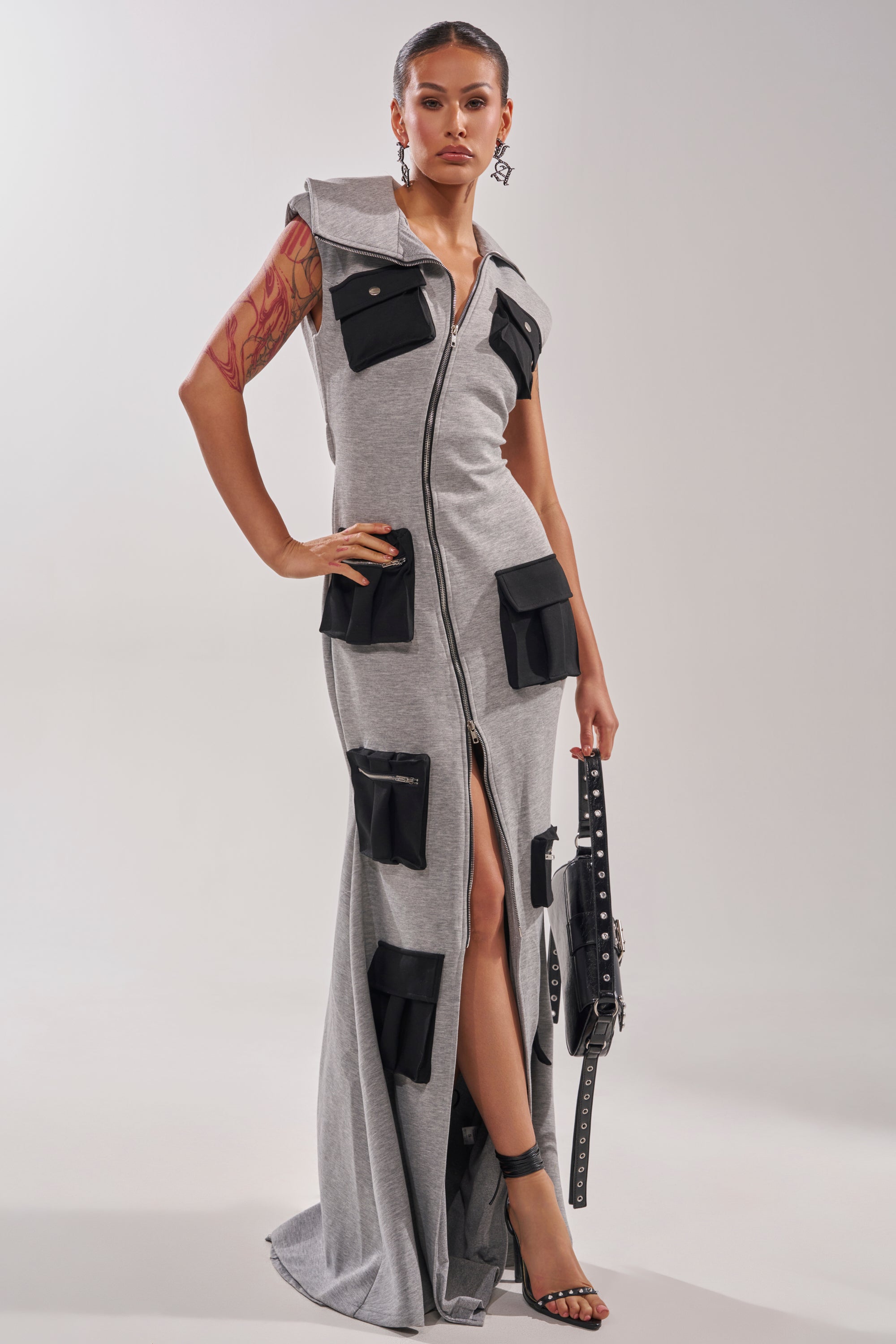 A woman stands confidently in the DON'T MESS WITH ME Collared Maxi Dress in Grey Scuba, featuring zip-front styling and cargo-inspired pockets. She carries a black studded handbag, wears black heels, and shows a tattoo on her left arm.
