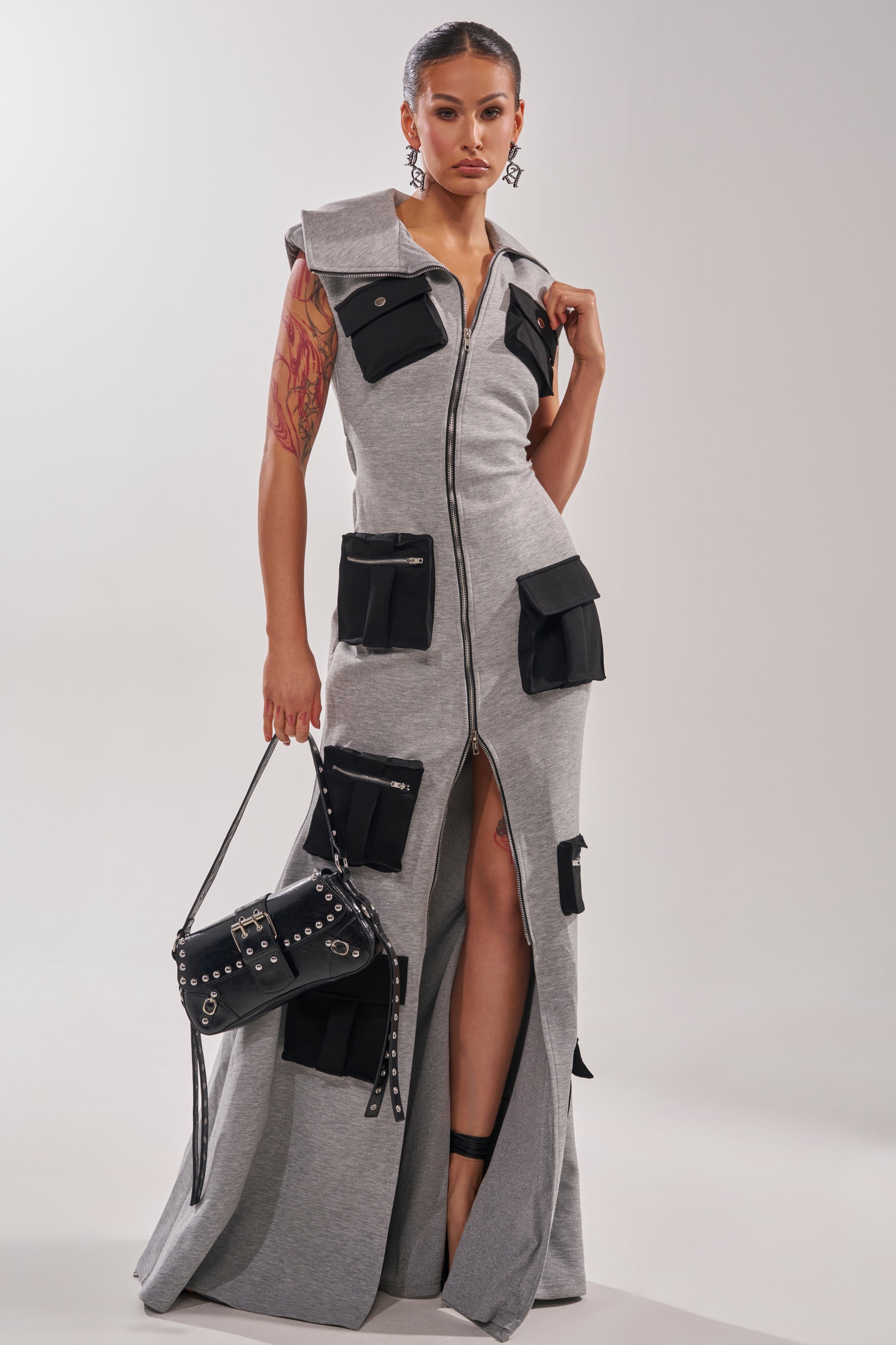 A woman wears the DON'T MESS WITH ME Collared Maxi Dress in Grey Scuba, featuring a sleeveless zip-front design, black patch pockets, and a high slit. She accessorizes with a black studded bag, earrings, and has her hair pulled back.