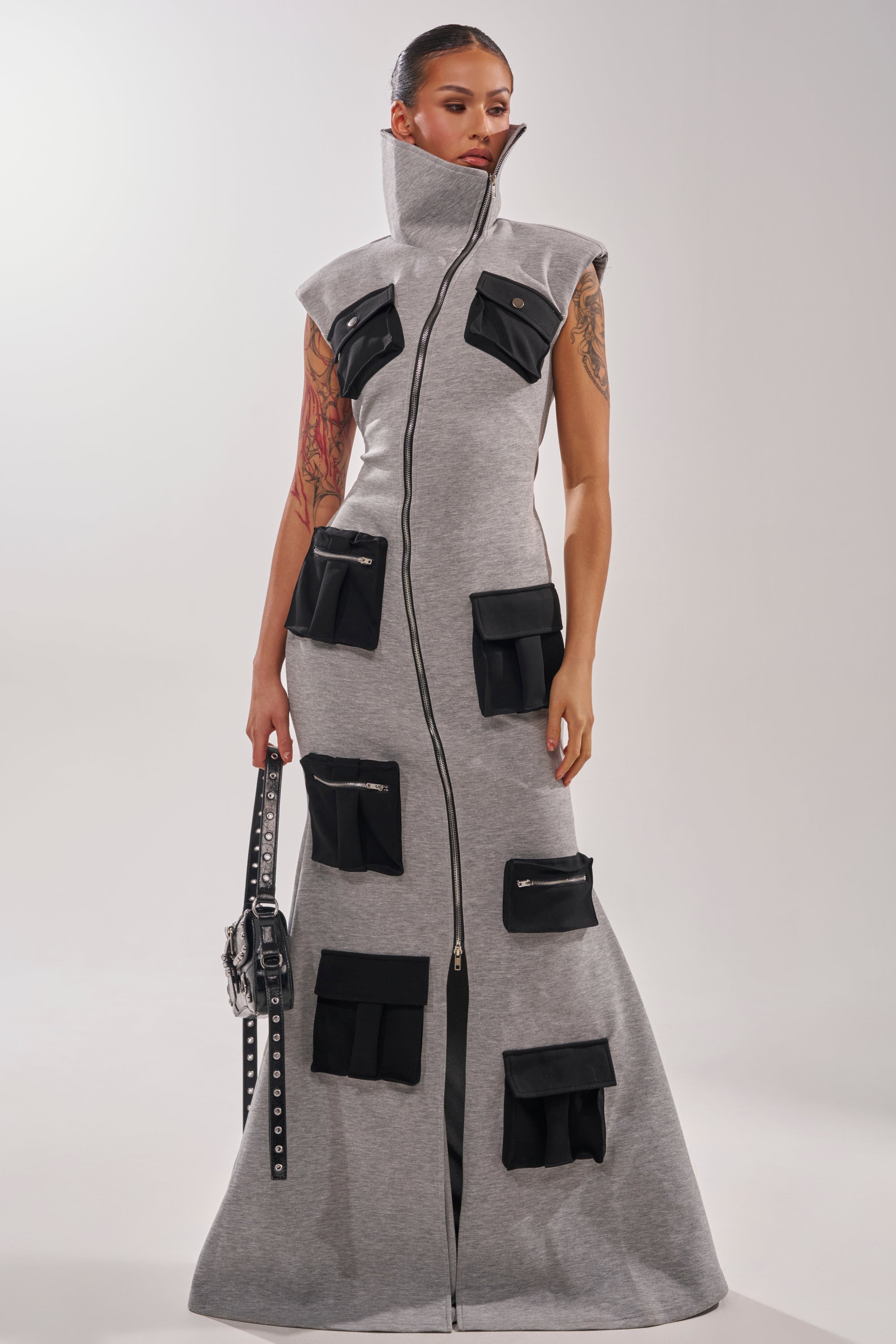 A woman stands confidently in the DON'T MESS WITH ME COLLARED MAXI DRESS IN GREY SCUBA, featuring oversized black cargo pockets. She carries a black studded handbag, her hair is sleek, and tattoos decorate her arms.