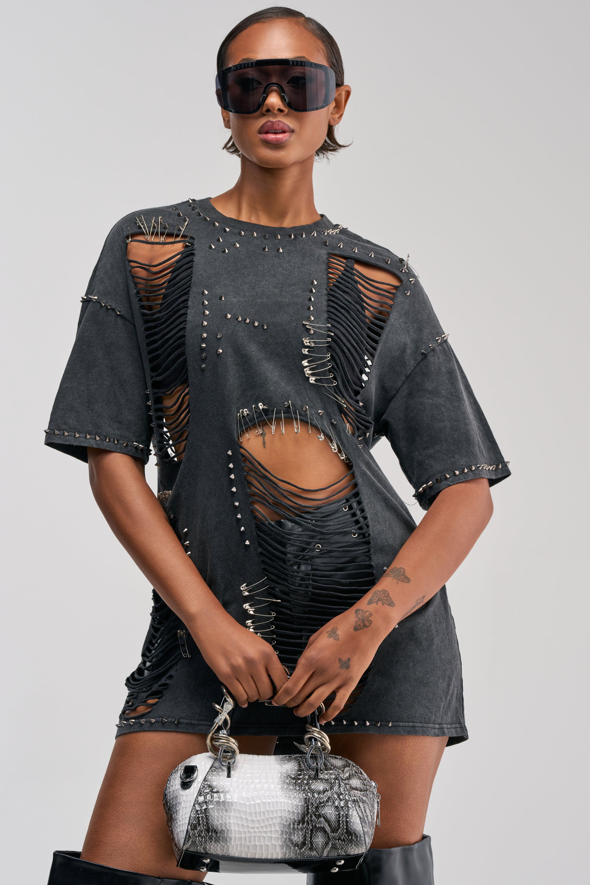 Wearing the MARGOT DISTRESSED STUDDED SHORT SLEEVE TEE styled with heavy distressing, metal studs, and safety pins, this person completes the look with large sunglasses, a silver handbag, and tall black boots while standing against a plain background.