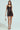 A woman models the SHOW US WHAT YOU GOT satin mini dress, showcasing its chocolate hue and lace trim bodycon fit. She accessorizes with a small rectangular metallic handbag and strappy high heels against a plain background.