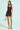 A woman wearing the "Show Us What You Got" satin mini dress, featuring black lace trim and strappy high heels, stands against a light background with a small transparent handbag. Her straight hair cascades over her shoulders as she confidently faces the camera.