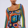 A woman with a confident expression wears the PARADISE FOUND KNIT MINI DRESS, displaying its vibrant crocheted design and long sleeves. The boho-chic mini dress features an array of bright colors and intricate patterns. She accessorizes with gold hoop earrings and stands against a plain background.